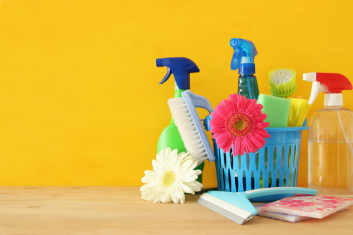 Cleaning supplies on a wood surface. There are two bright colored daisies with the cleaning products. They are all lined up in front of a blank yellow background