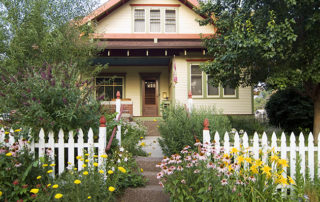 One story home with a white picket fence in the ground. The front yard is full of spring blooms and newly-leafing trees.