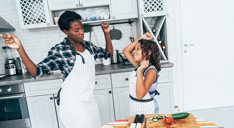 Mother and daughter dancing while cooking in the kitchen. Mother is Black, daughter is mixed race.