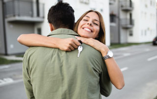 Happy young couple embracing. Young woman facing the camera over her partner's shoulder with the keys in her hand in front of their new home.