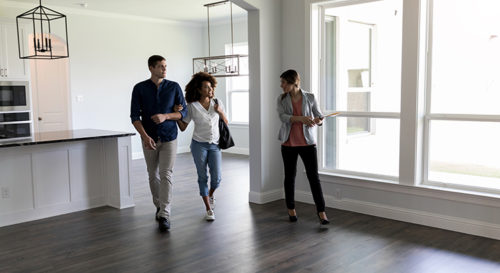 Young couple meeting a realtor inside a property. They're walking arm in arm in an empty living room, right by a large window
