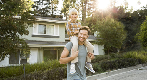 young boy sitting on father's shoulders, both smiling, standing outside a house at sunset.