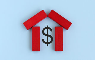 wooden blocks laid on a flat background. The block are arranged in a basic house shape, and are surrounding a dollar sign in the middle.