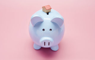 light blue piggy bank on a pastel pink background. there is a tiny wooden house model on top of the piggy bank