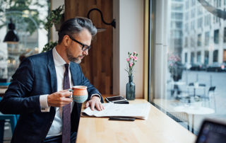 Mature businessman reading paper while having coffee at a coffee shop. He is facing a window looking out onto the street
