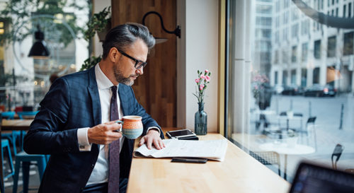 Mature businessman reading paper while having coffee at a coffee shop. He is facing a window looking out onto the street