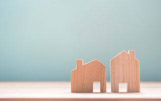two wooden blocks shaped like different sized homes. Set on a flat wooden surface, and a blank blue paper background